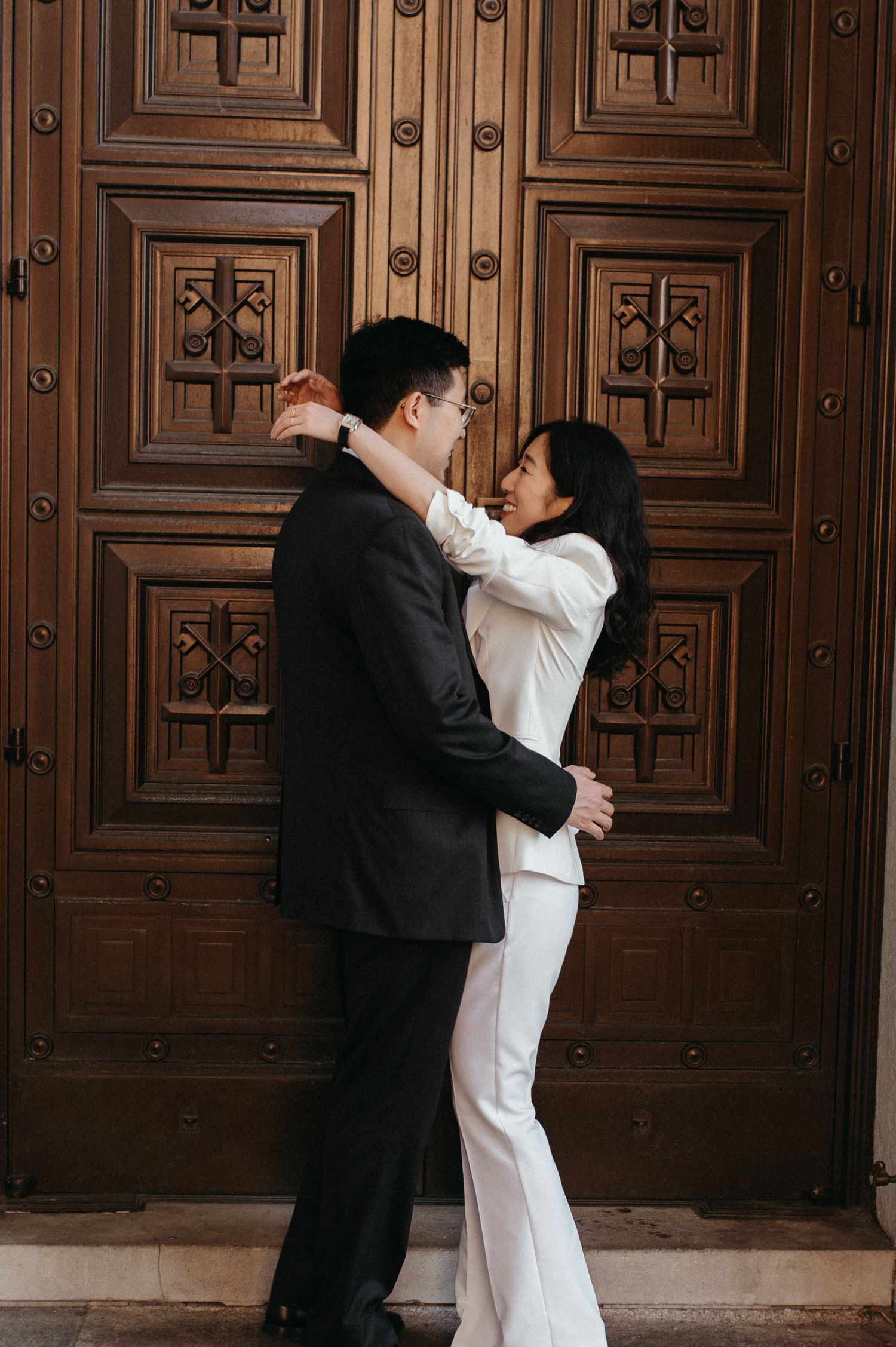 A couple in contrasting black and white outfits share a romantic moment against decorative wooden doors.