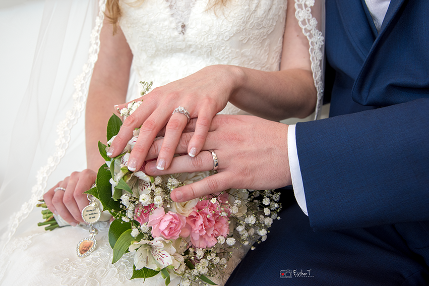 Newlywed couple showing wedding rings resting on pink and white bridal bouquet.