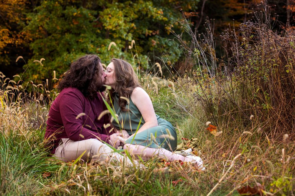 Couple sitting and kissing in a meadow surrounded by tall grass and autumn foliage.