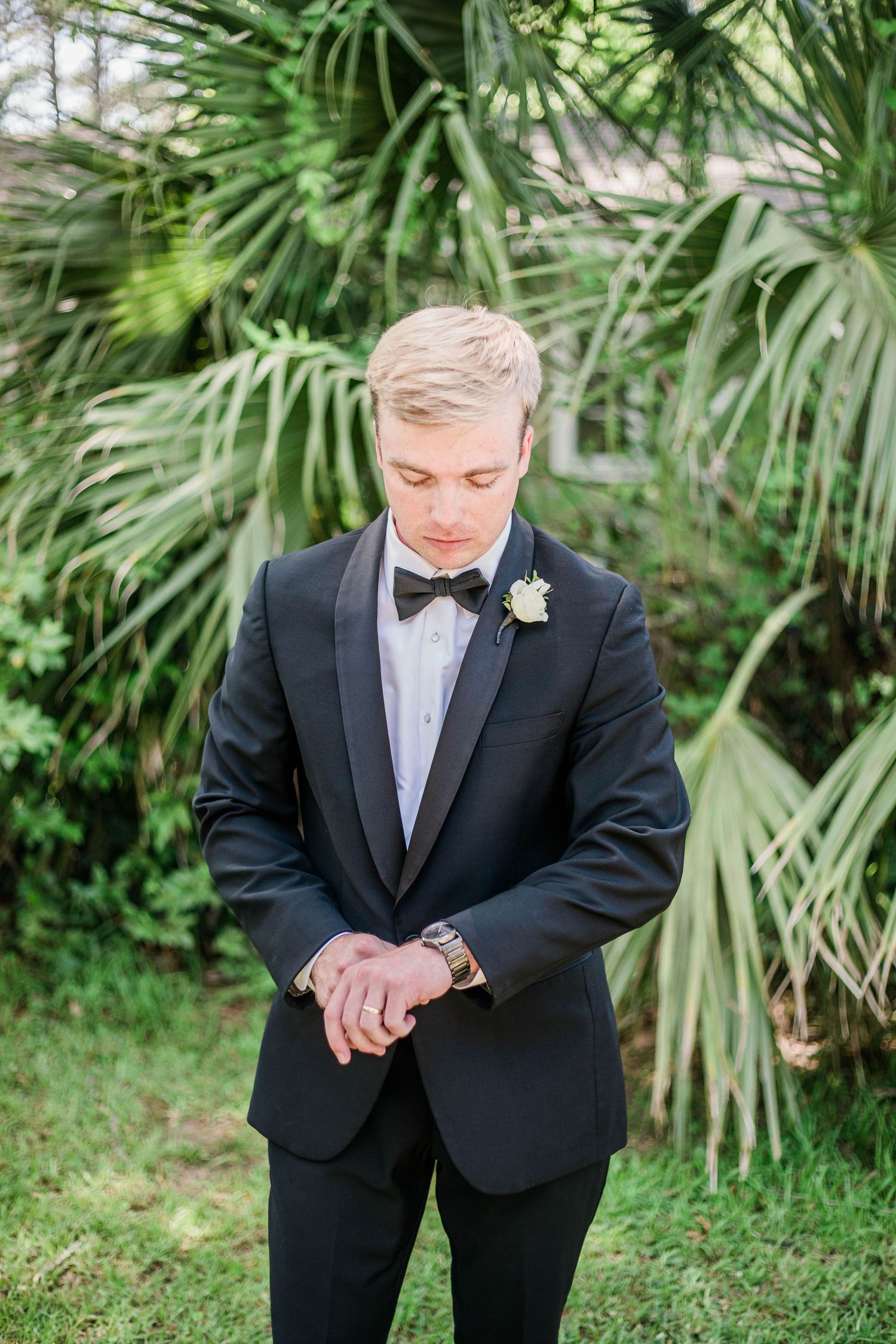 Groom in classic black tuxedo checks his watch while standing in front of palm trees.