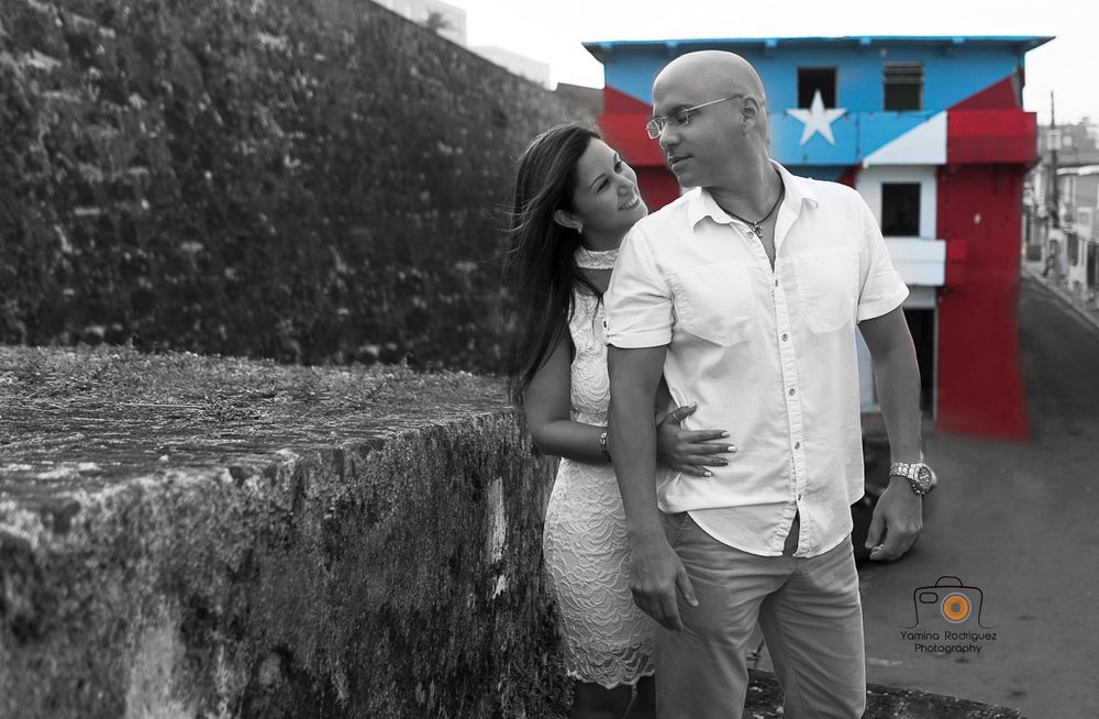 A couple embraces against a weathered wall with a Puerto Rican flag in the background.