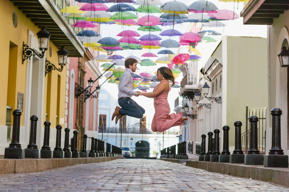 A joyful couple leaps beneath colorful suspended umbrellas on a charming colonial street.