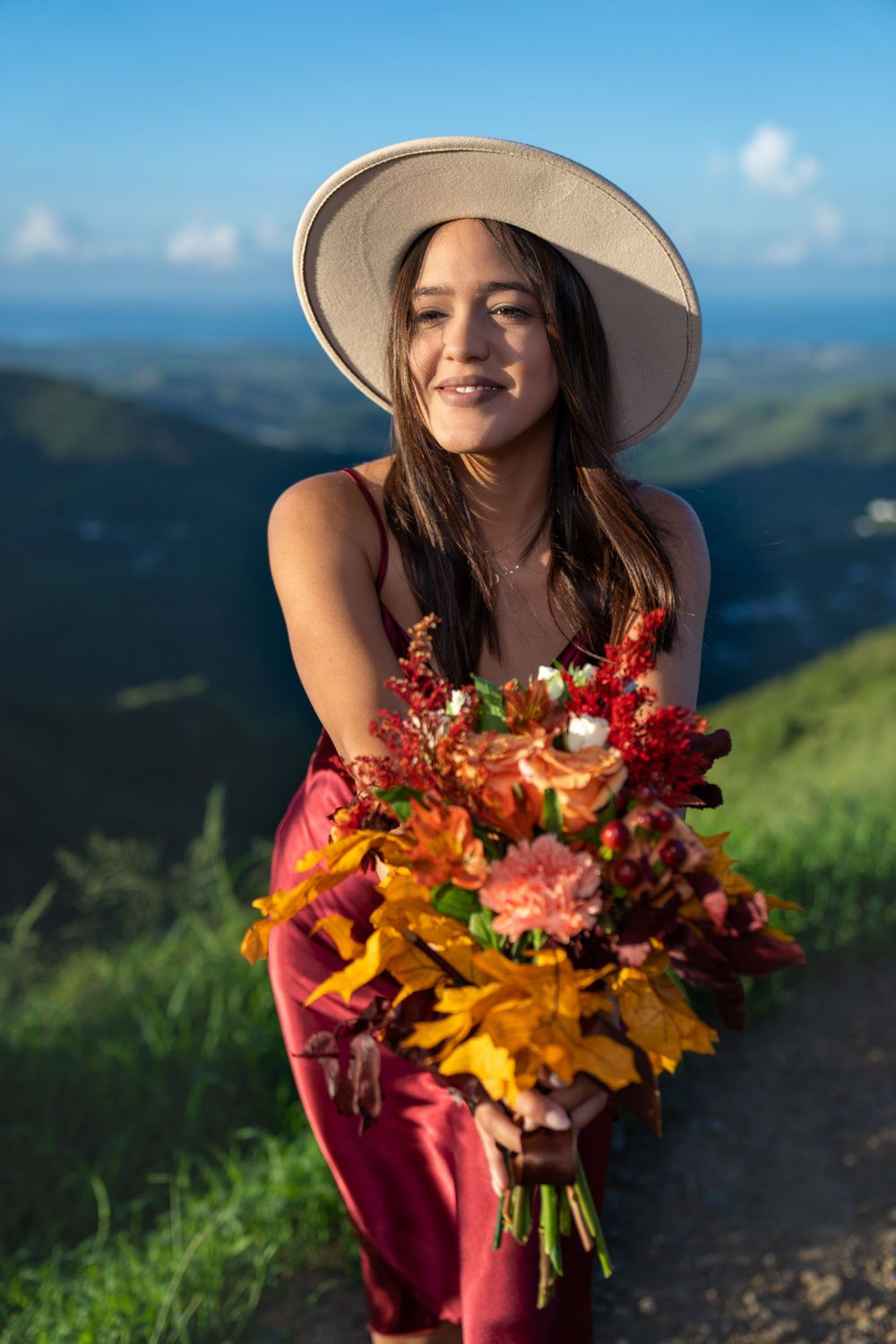 A person in a red dress and hat holding a vibrant autumn bouquet against a mountainous backdrop.