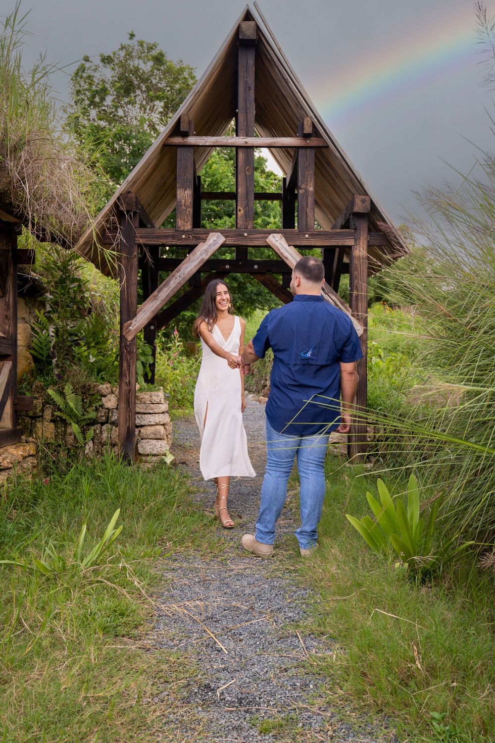 A couple standing on a path near a rustic A-frame structure with a rainbow visible in the stormy sky.