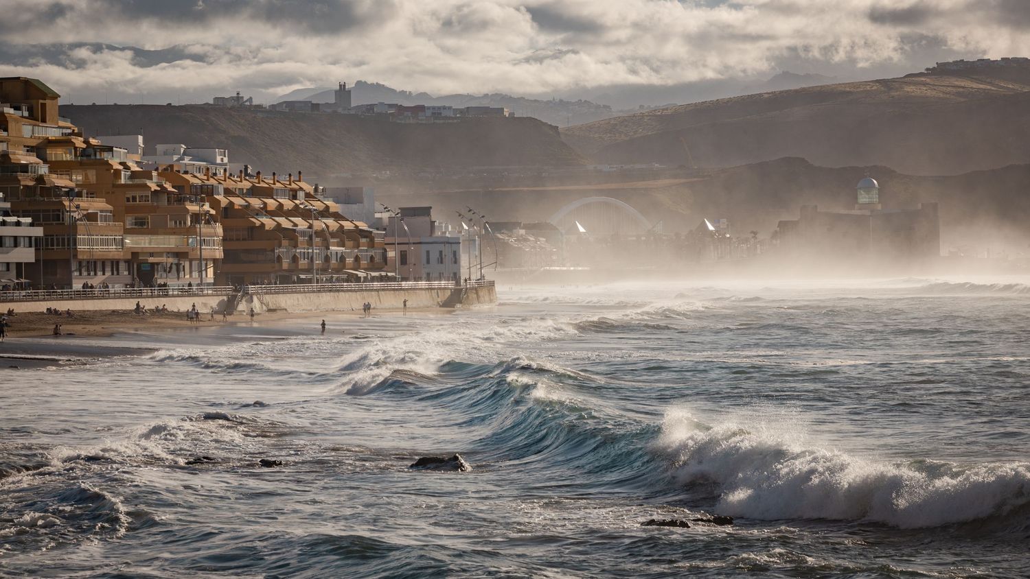Stormy coastal town with crashing waves and misty sea spray along a rugged shoreline under dramatic cloudy skies.