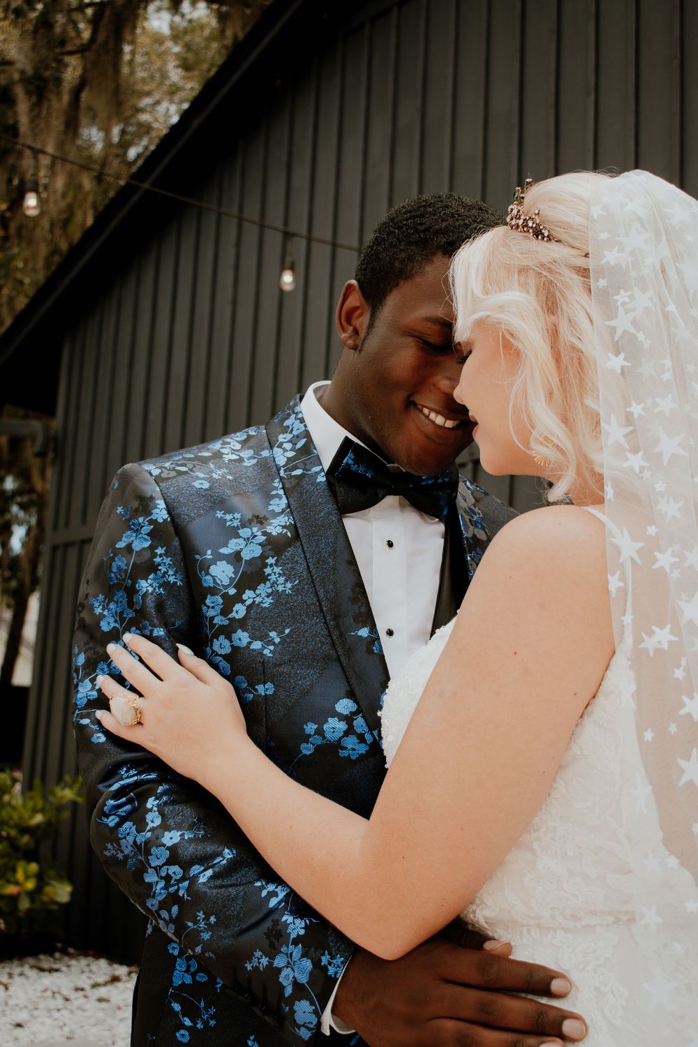 Newlyweds embrace in formal wedding attire, featuring a floral patterned suit and star-detailed veil.