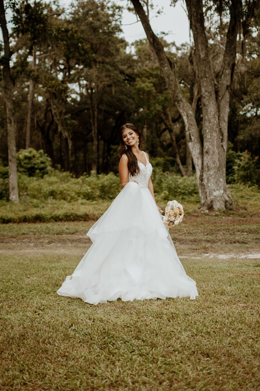 Bride in flowing white wedding dress twirls gracefully on grassy field surrounded by trees.