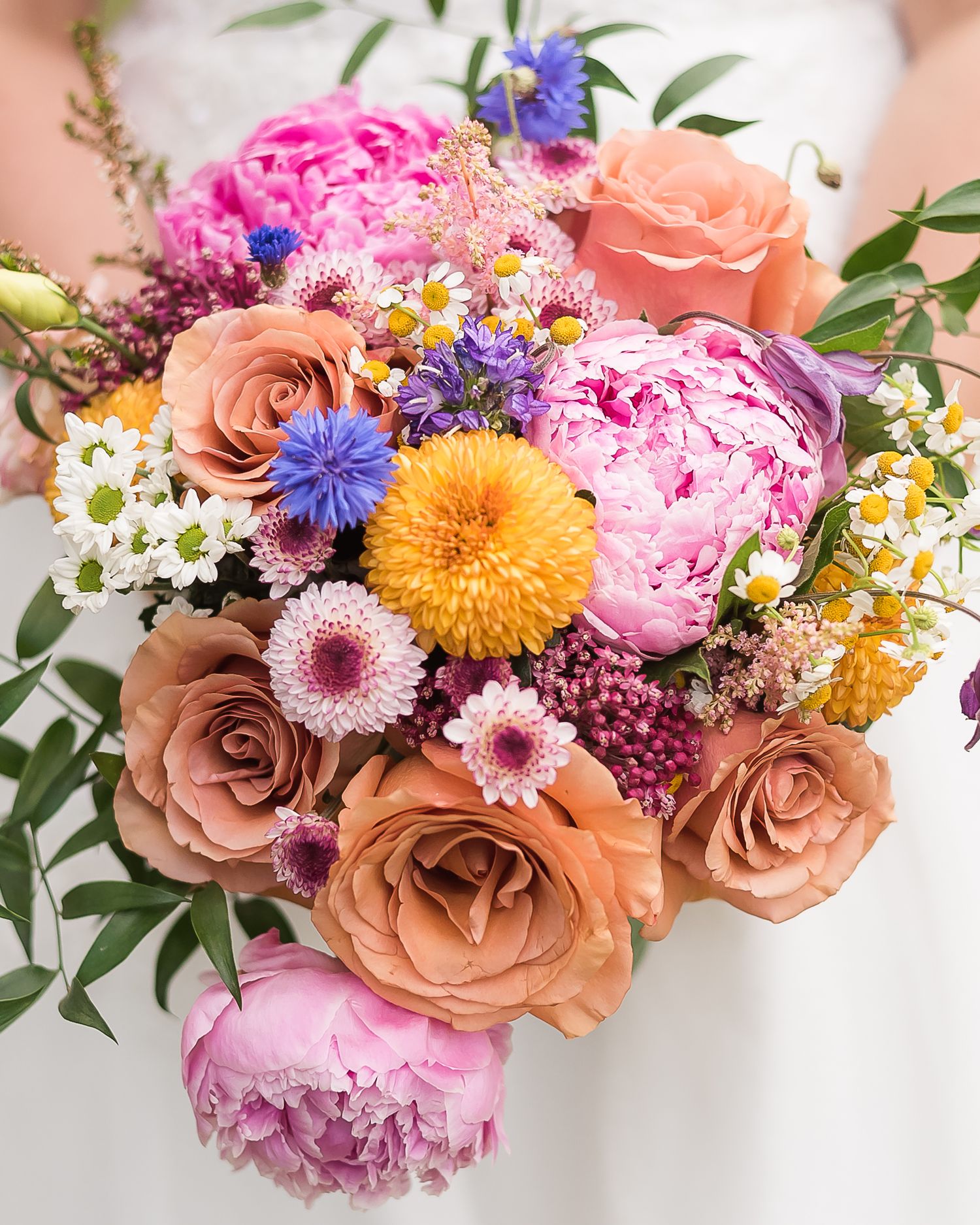 A close up image of a gorgeous, colourful, bridal bouquet. Pink flowers, orange roses, peonies, daisies, greenery.