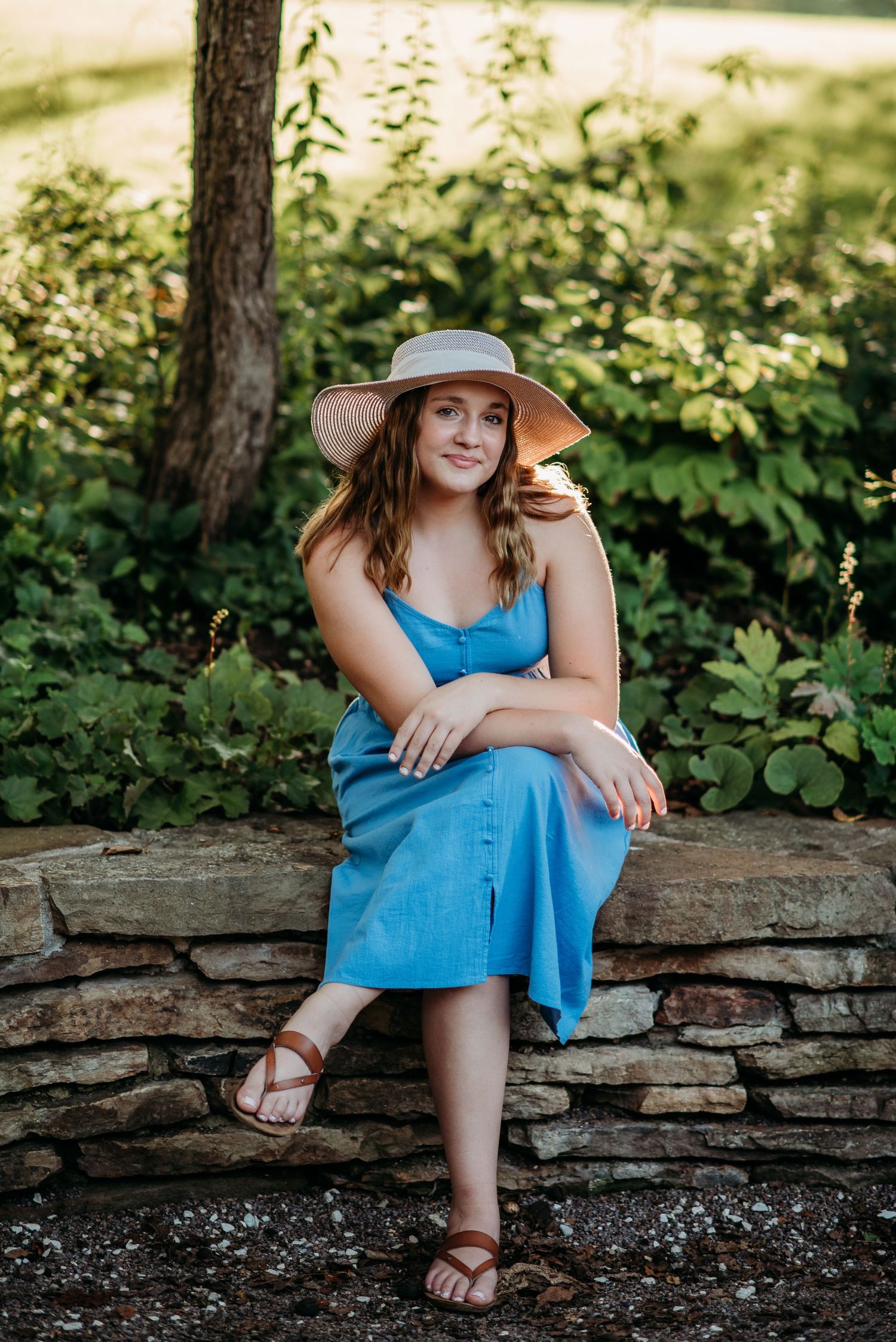 Person in blue dress and sun hat sitting casually on stone wall outdoors.