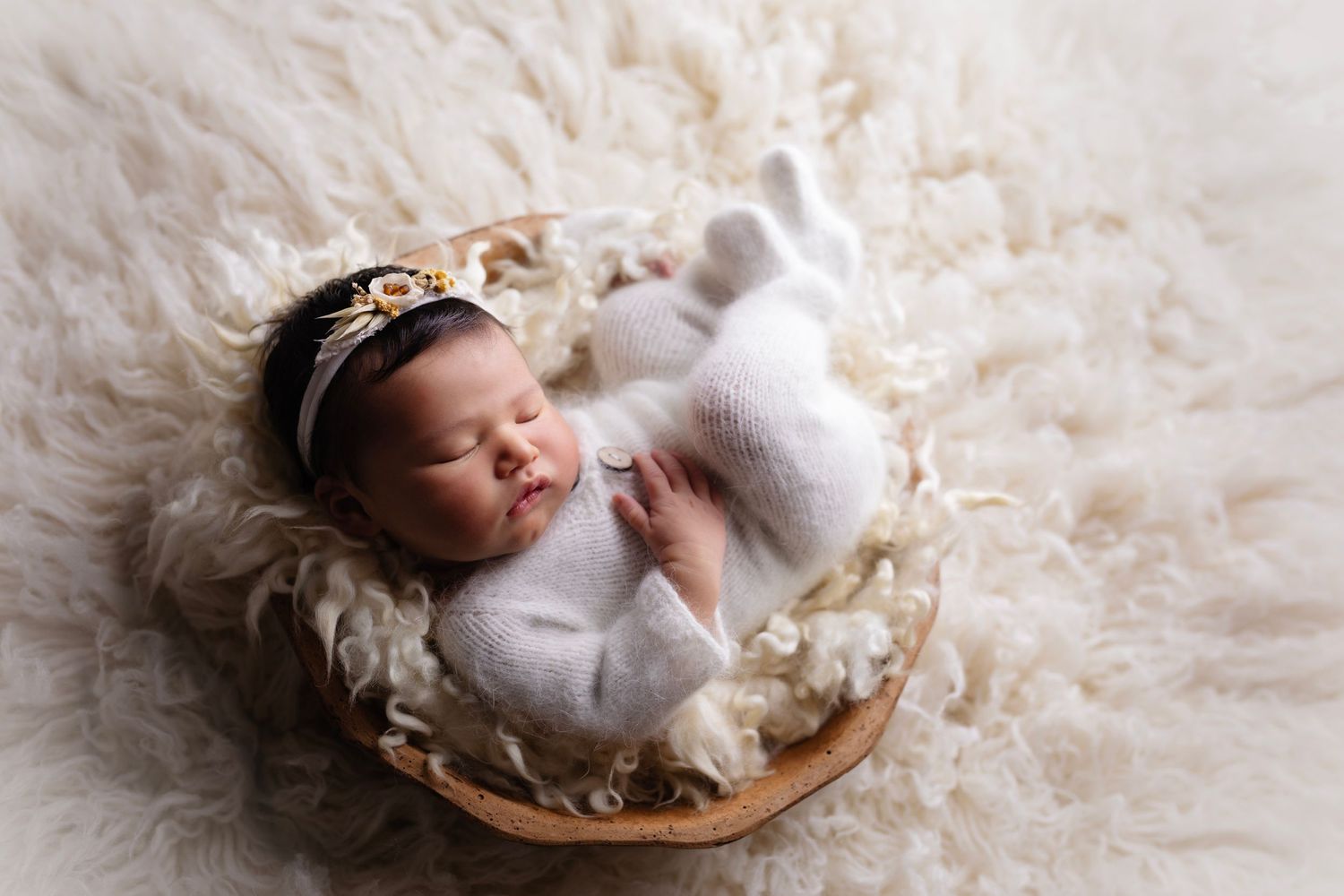 Newborn baby sleeping peacefully in basket on cream fluffy blanket wearing white outfit and crown.