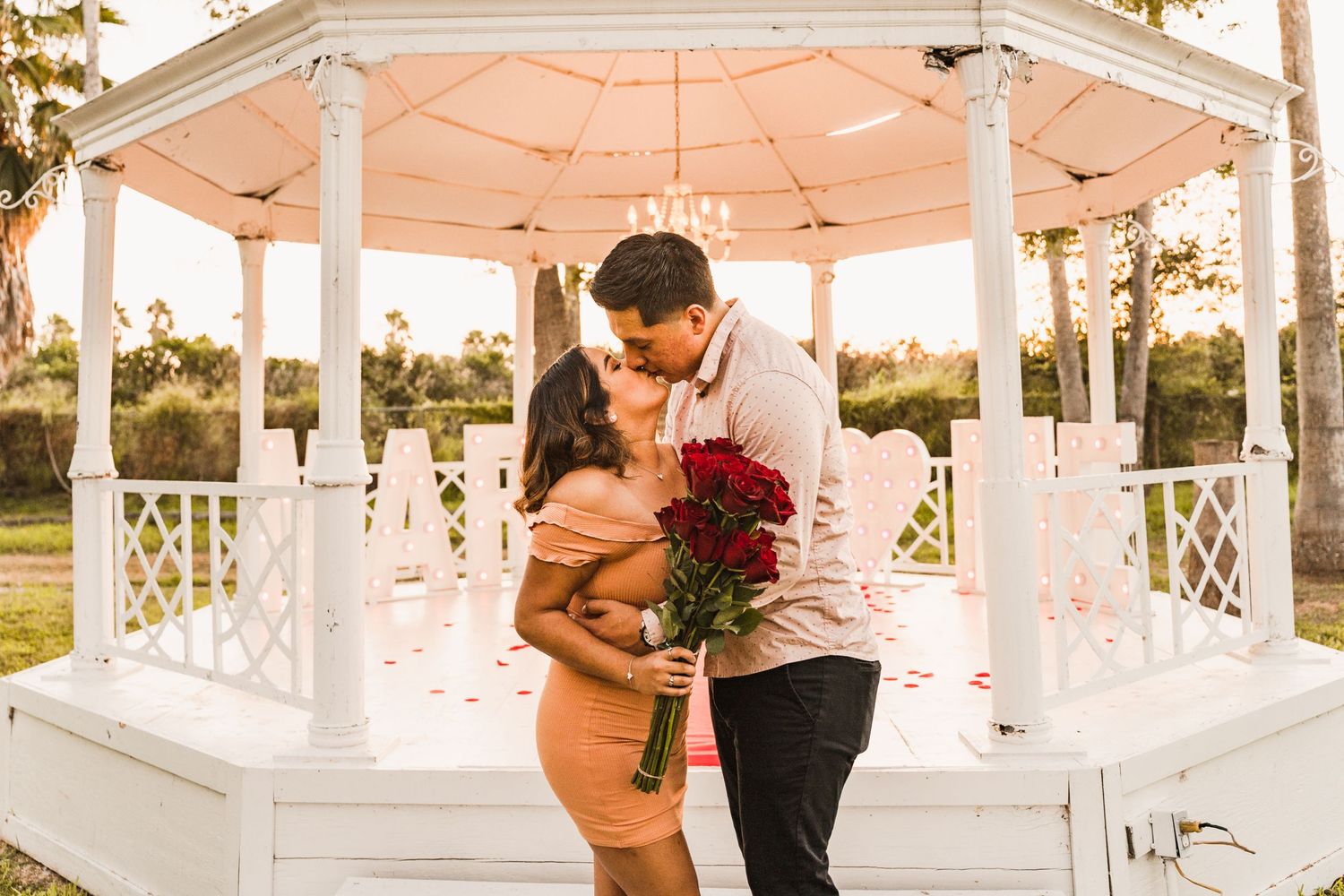 Couple sharing kiss under white gazebo with red roses during romantic outdoor proposal.