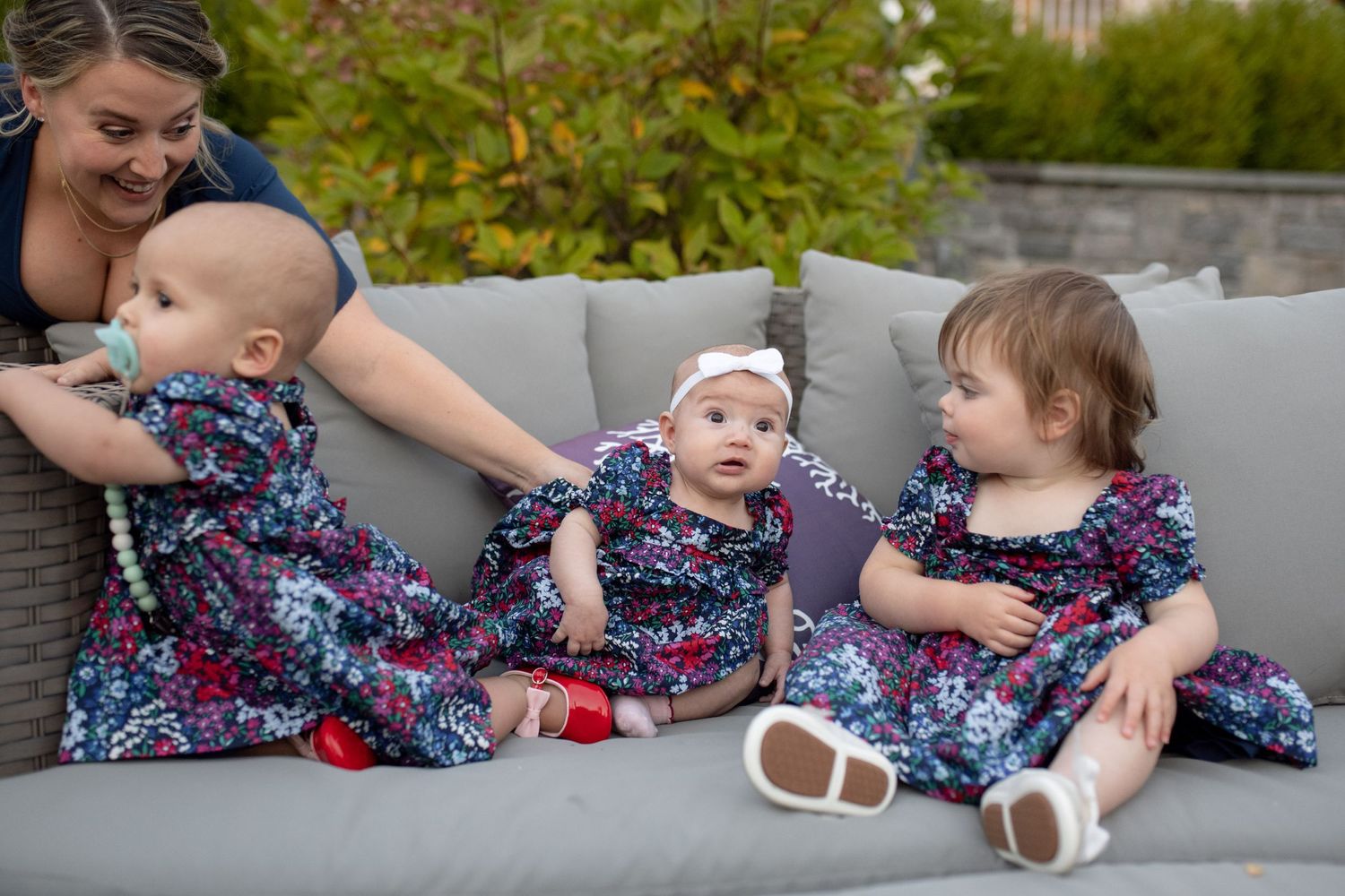 Three babies wearing matching floral dresses sitting on grey outdoor furniture.