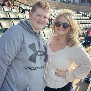 Teenage boy in Under Armour hoodie posing with smiling blonde woman at an outdoor sports stadium.
