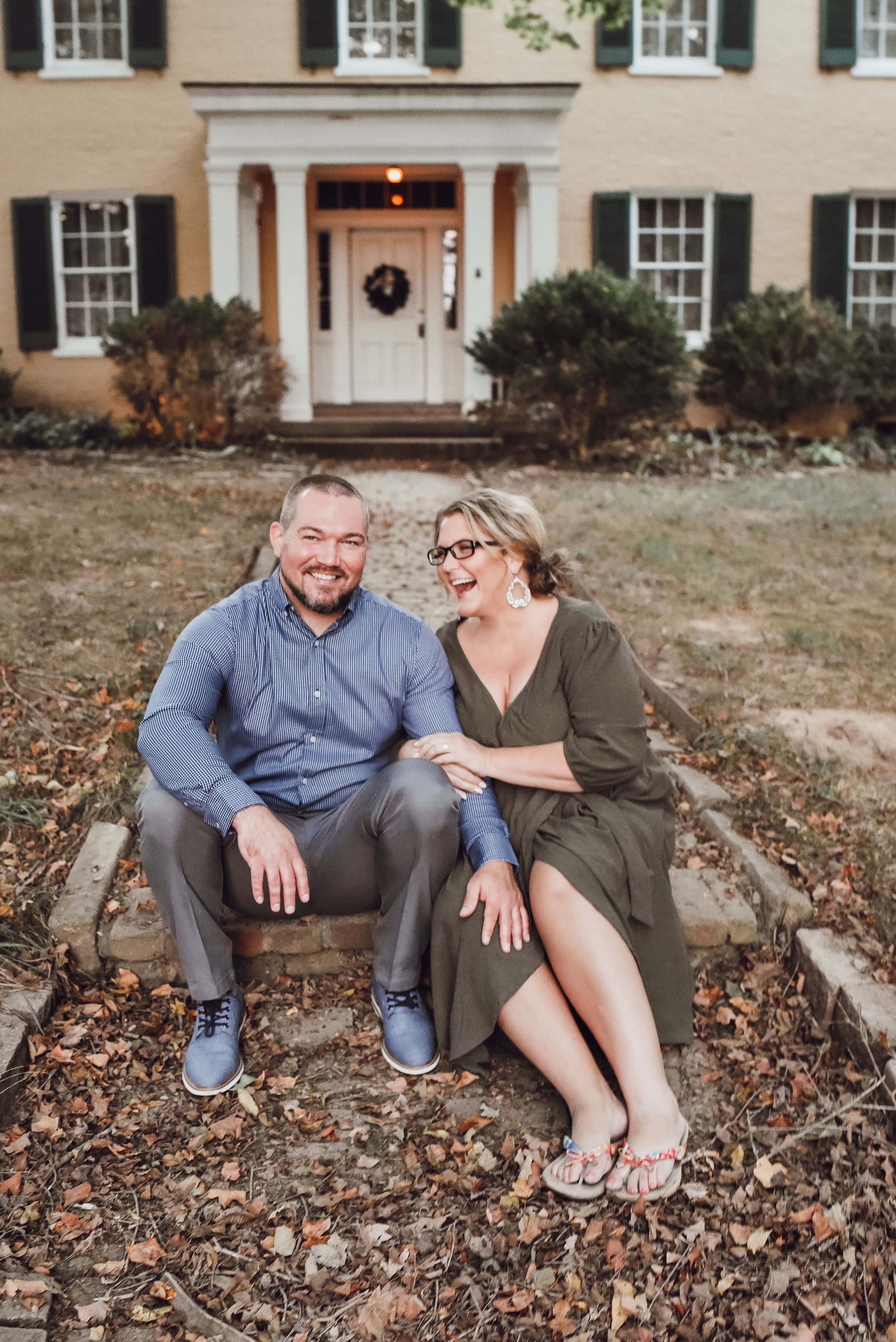 Smiling couple sitting together on stone steps in front of a classic yellow colonial home surrounded by fall leaves.