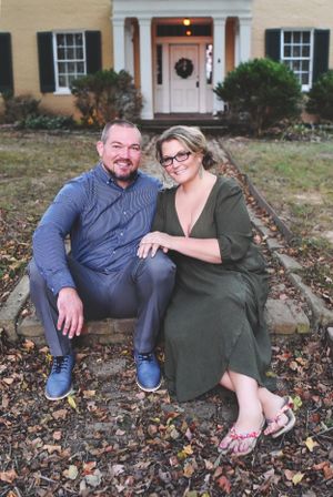 Happy couple posing and smiling together on stone steps outside a traditional yellow colonial home in autumn.