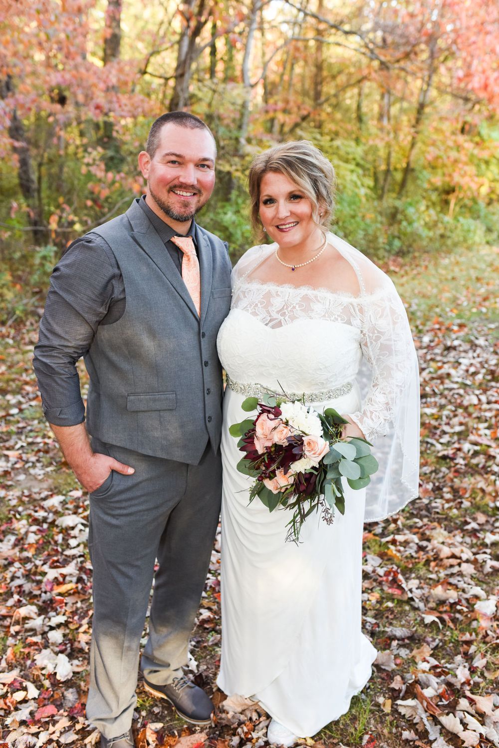 Smiling bride and groom standing among colorful fall leaves in forest, bride in lace gown holding bouquet.
