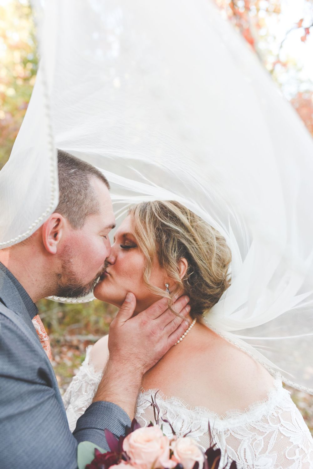 Bride and groom sharing romantic kiss under flowing white veil outdoors during fall wedding ceremony.