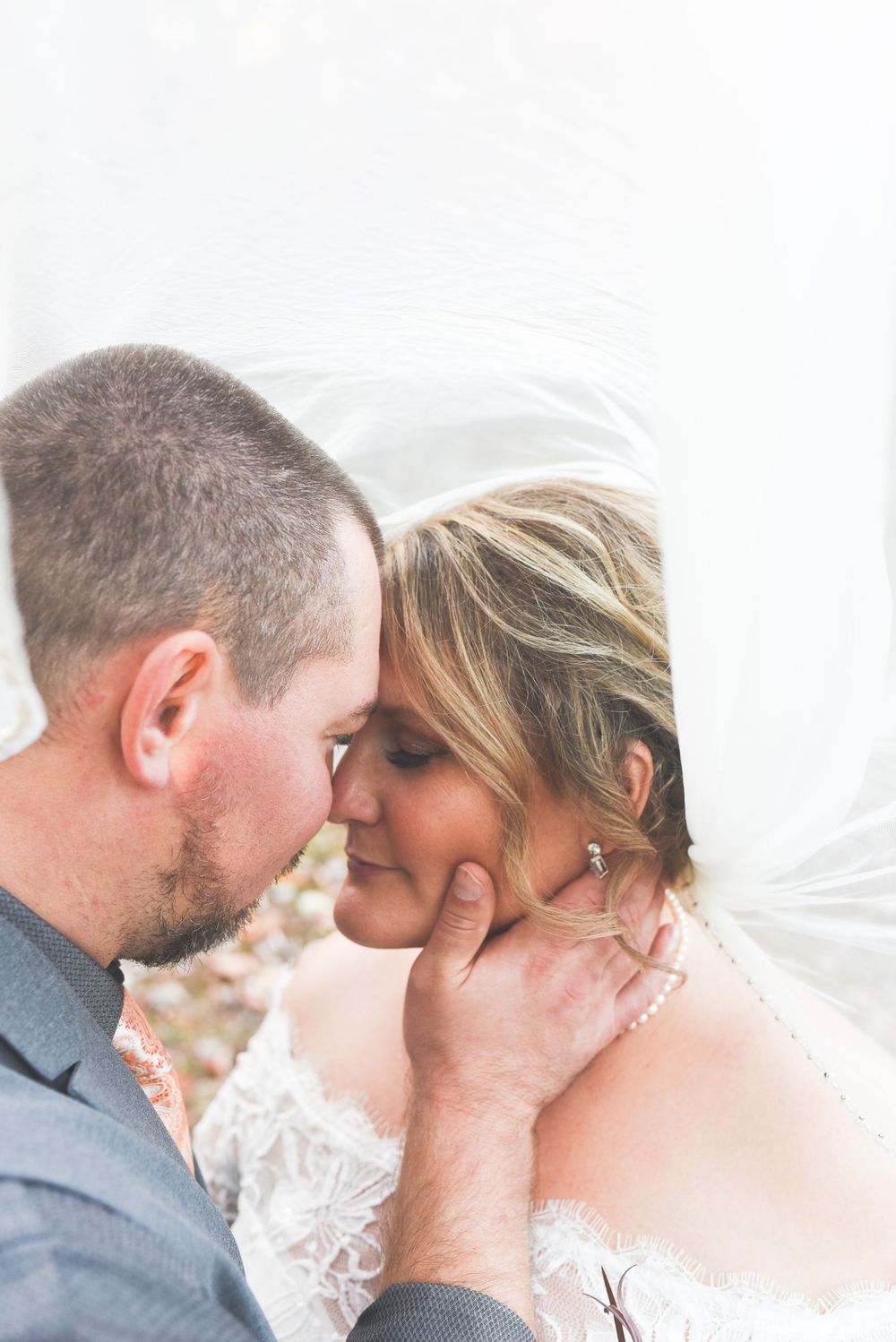Groom tenderly holding bride's face under wedding veil in intimate romantic outdoor autumn portrait.