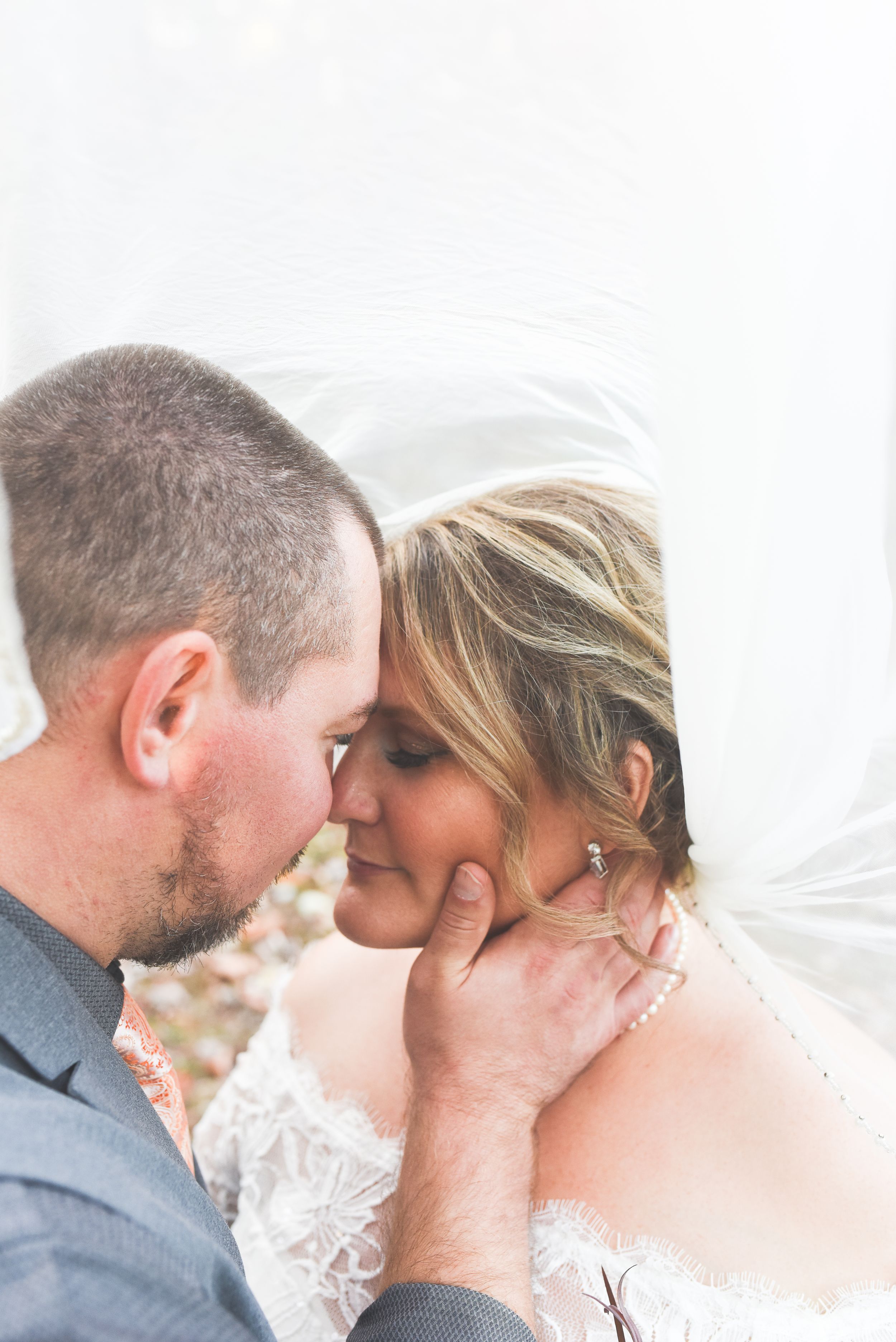 Groom tenderly holding bride's face under wedding veil in intimate romantic outdoor autumn portrait.