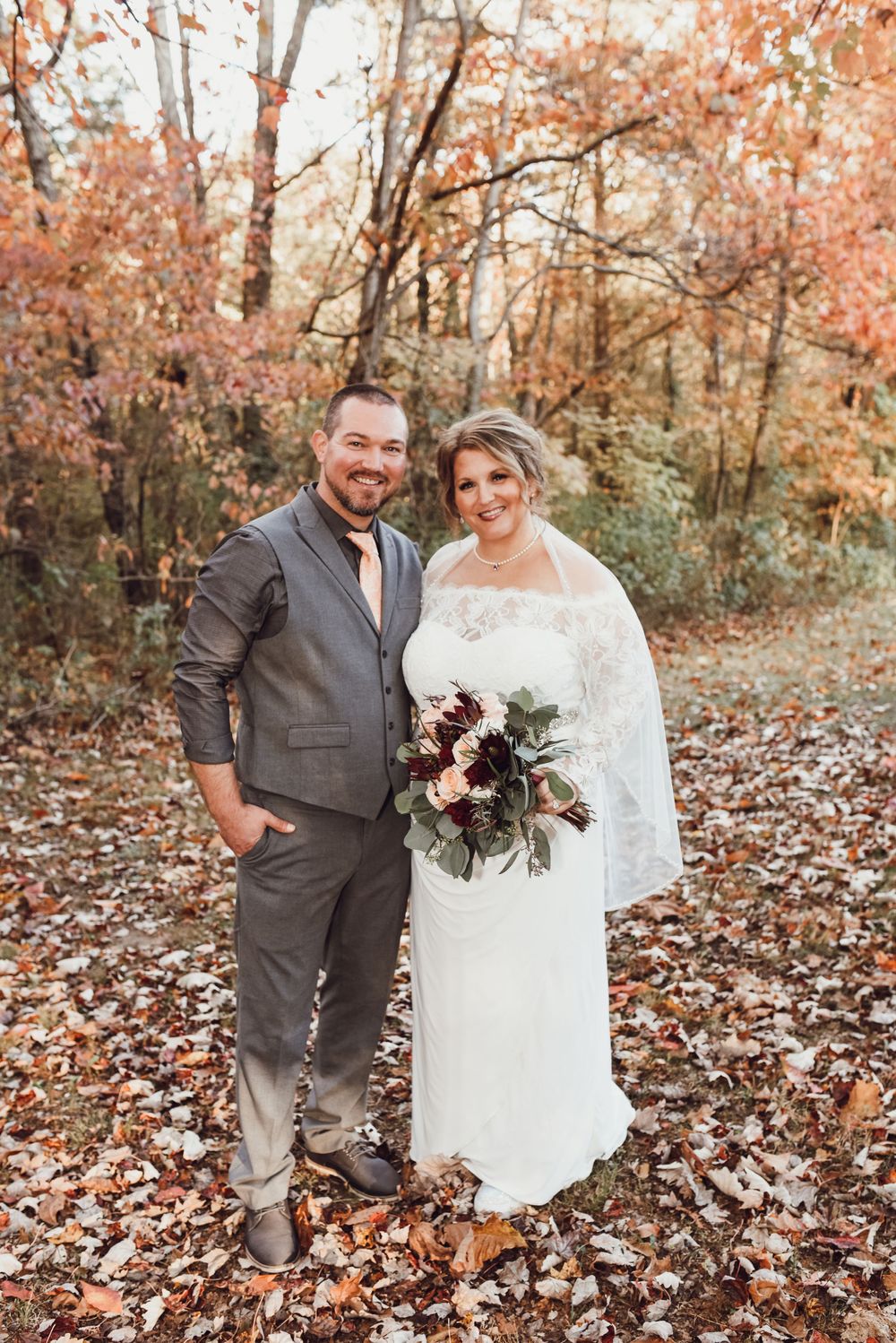 Bride and groom posing together in autumn forest with fall foliage, bride holding bouquet in lace wedding dress with veil.