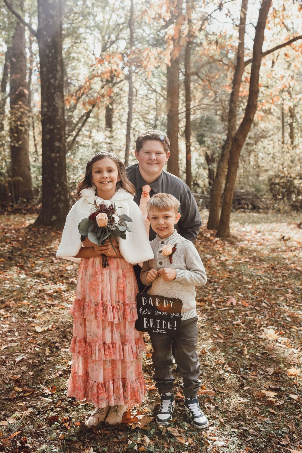 Three children posing in autumn woods, girl holding flowers, boy with Daddy here comes your bride sign.