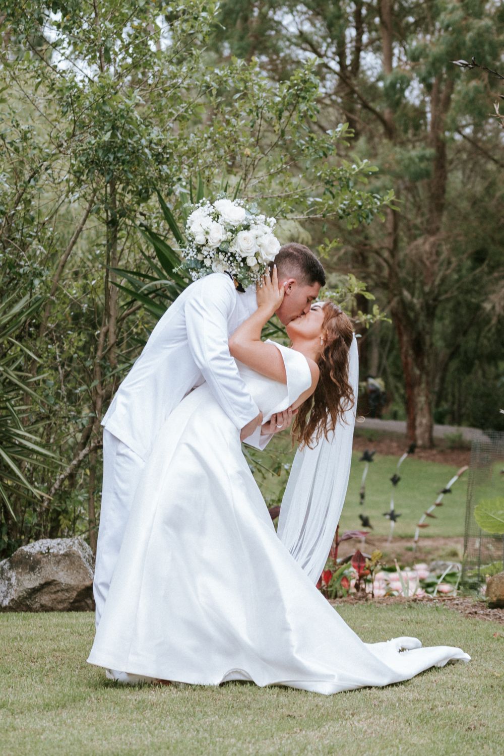 A romantic wedding couple in white sharing a dramatic dip kiss in a garden setting.