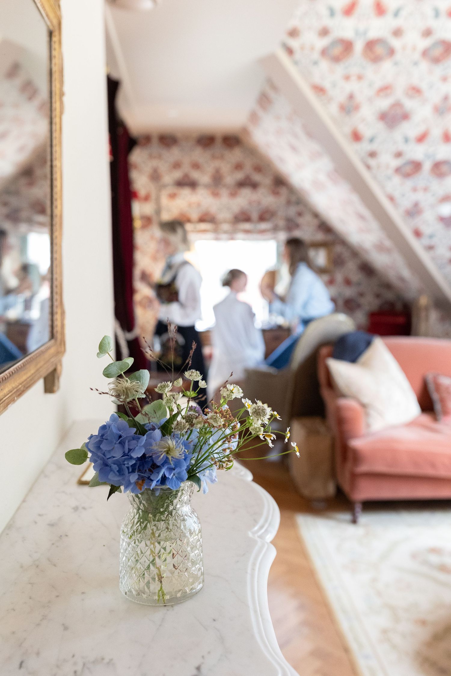 A blue hydrangea flower arrangement in a glass vase with floral wallpaper and pink couch in background.