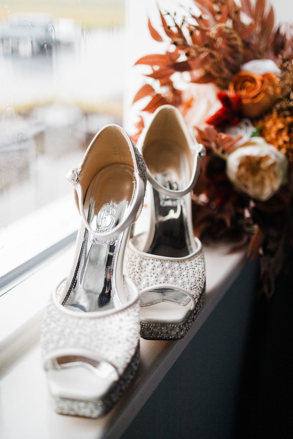 Sparkling silver wedding heels displayed next to autumn flowers on a windowsill.