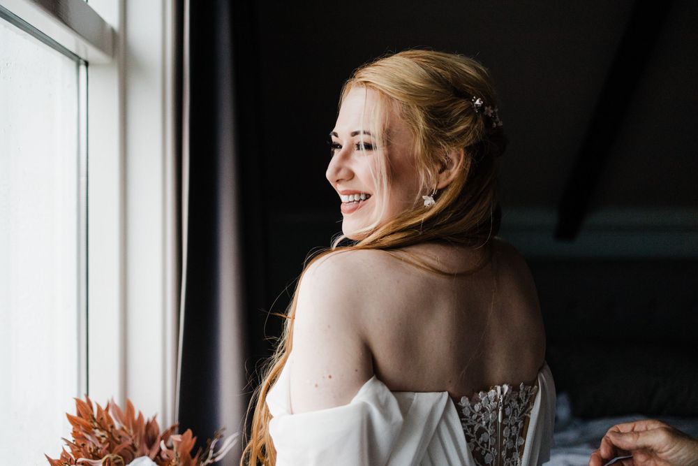 Person in white wedding dress smiling while looking out window with soft natural light.