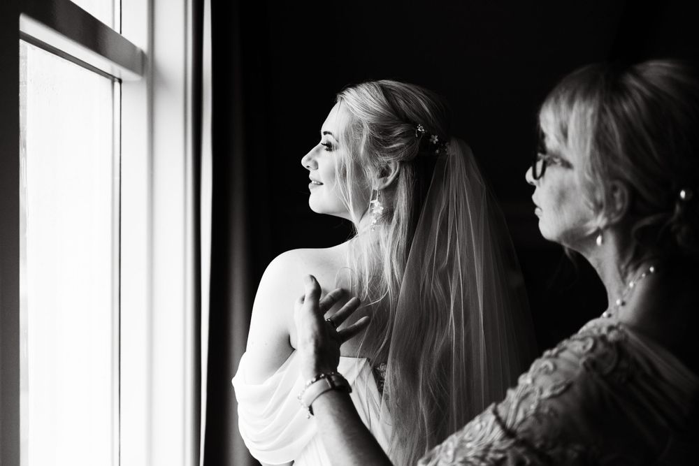Black and white photo of two people looking out window during wedding preparations.