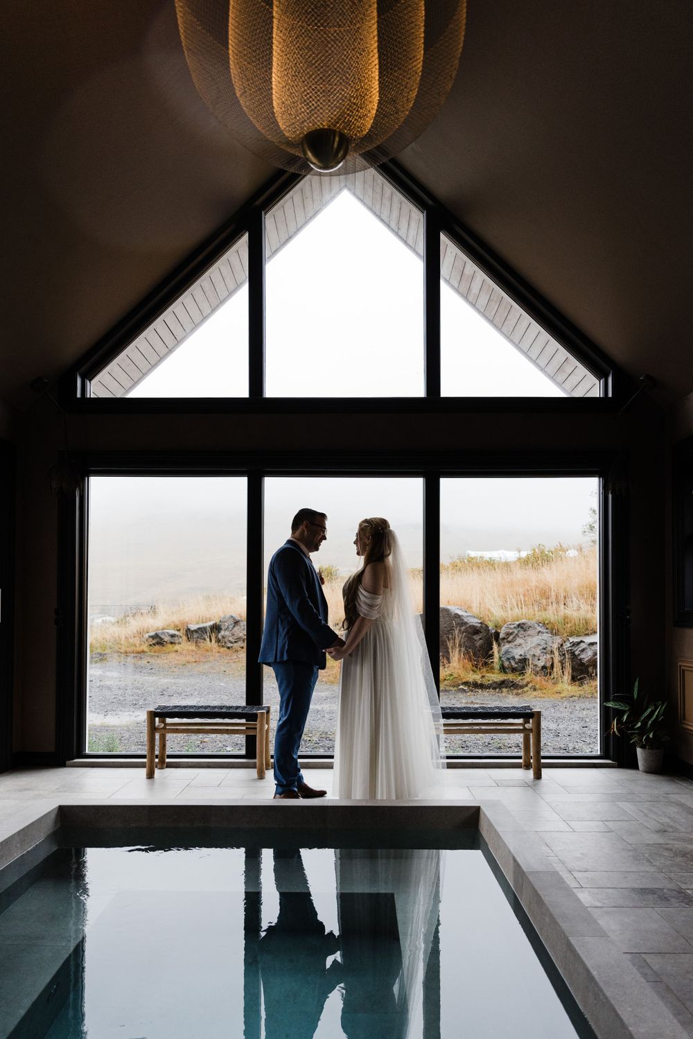 Couple in wedding attire silhouetted against large triangular window overlooking scenic mountain landscape.