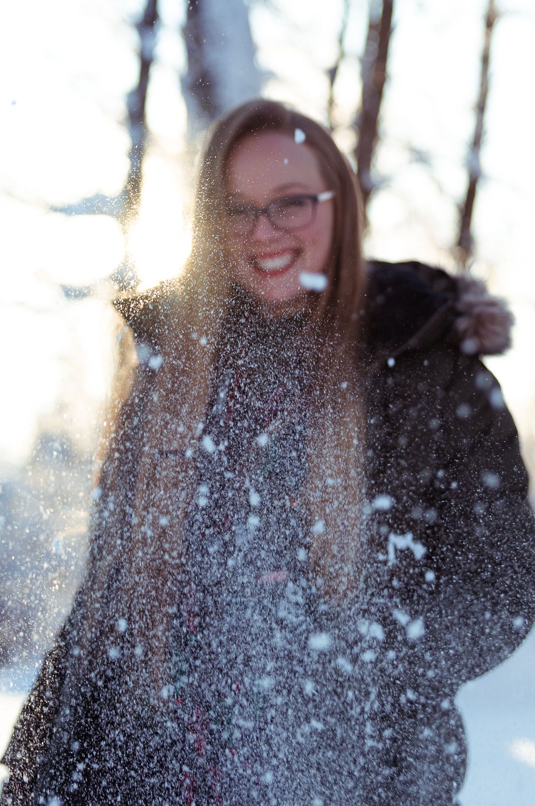 Person wearing glasses playfully enjoys falling snow in winter forest with sunlight streaming through trees.