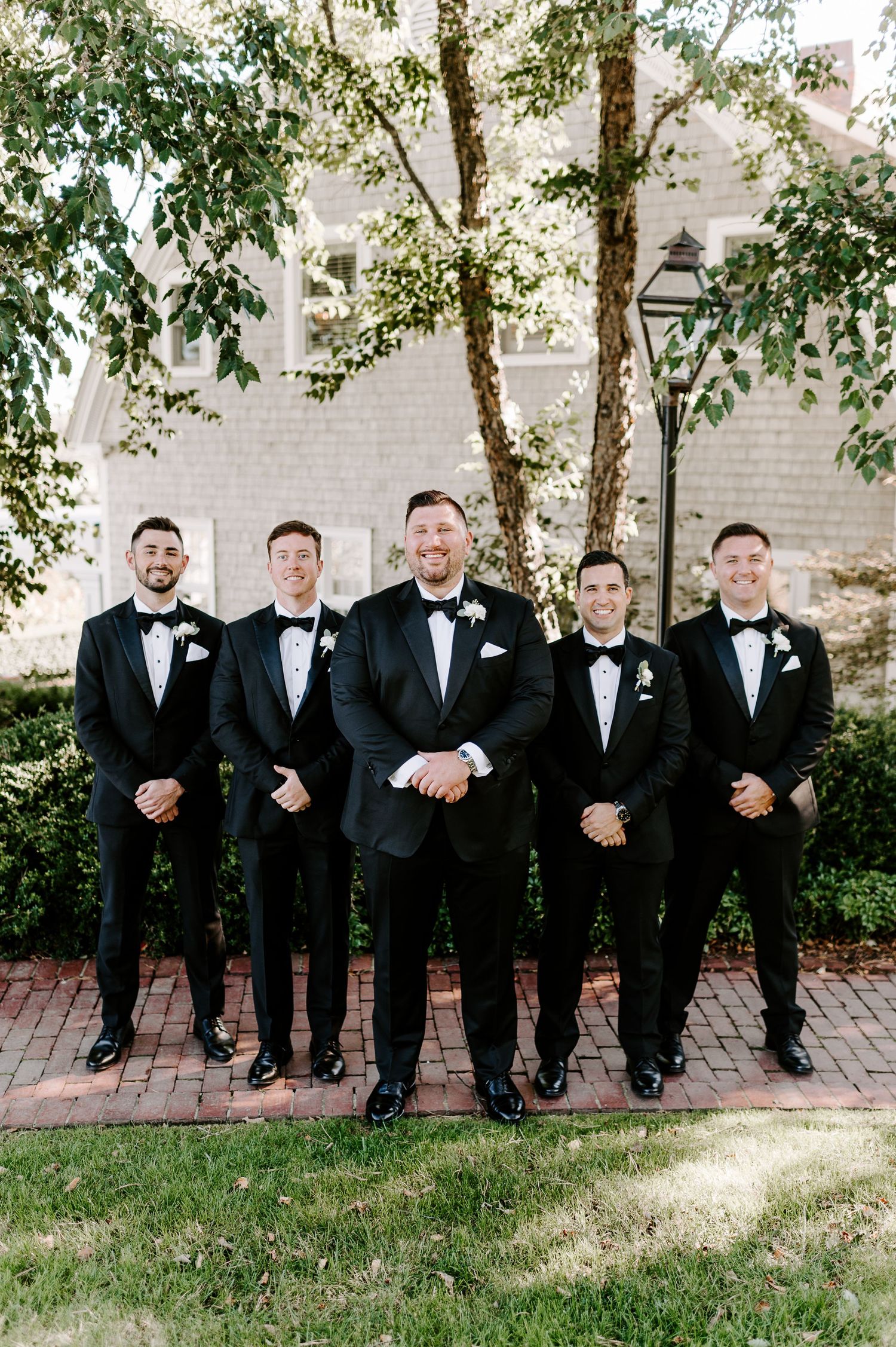 Groomsmen in black tuxedos sit casually on brick path for wedding photos.