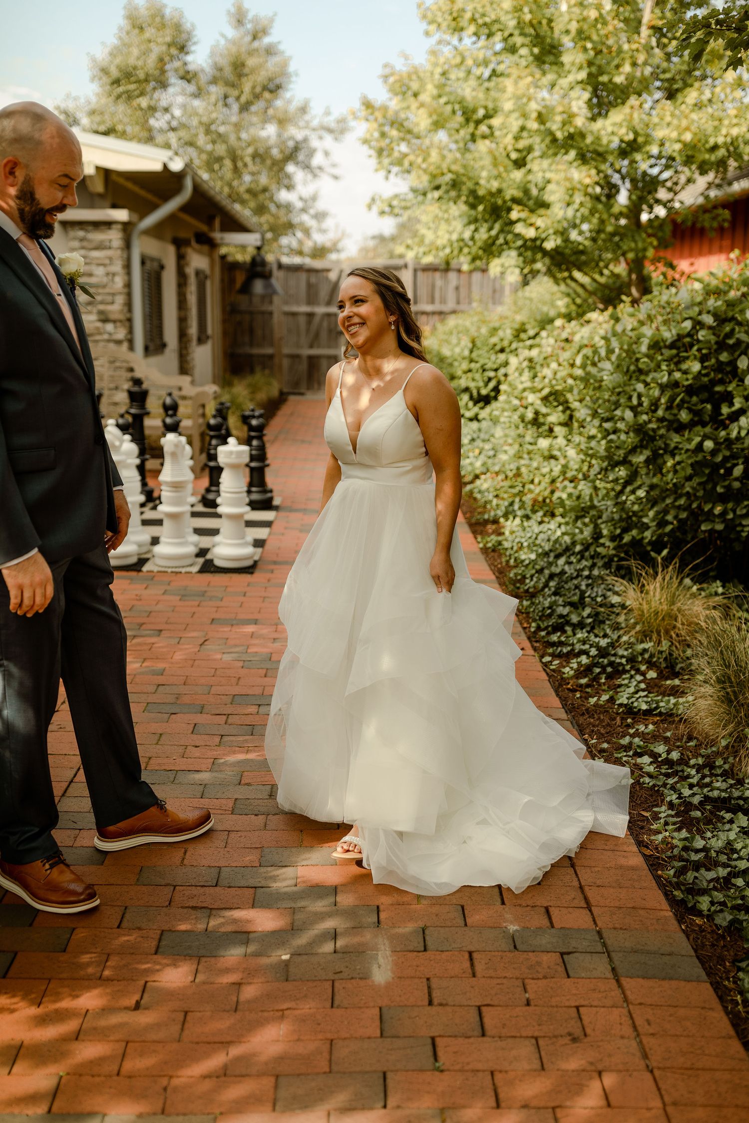 White wedding dress flowing across red brick patio with garden backdrop on sunny day.