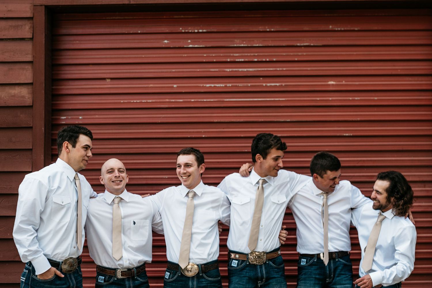 Groomsmen in casual white shirts and jeans pose casually against red barn siding.