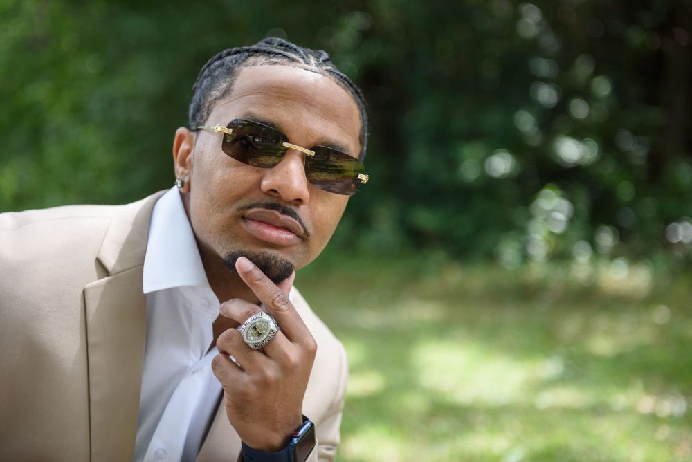 Man in beige suit jacket and sunglasses poses thoughtfully with hand on chin in outdoor setting.