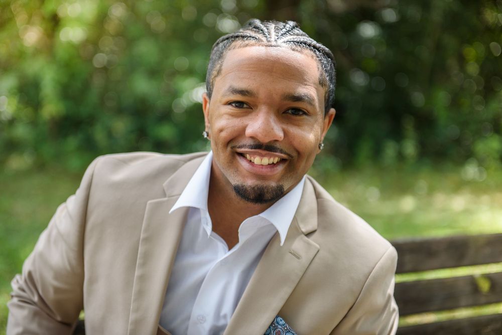Professional portrait of person wearing tan suit jacket and white dress shirt outdoors against blurred natural background.