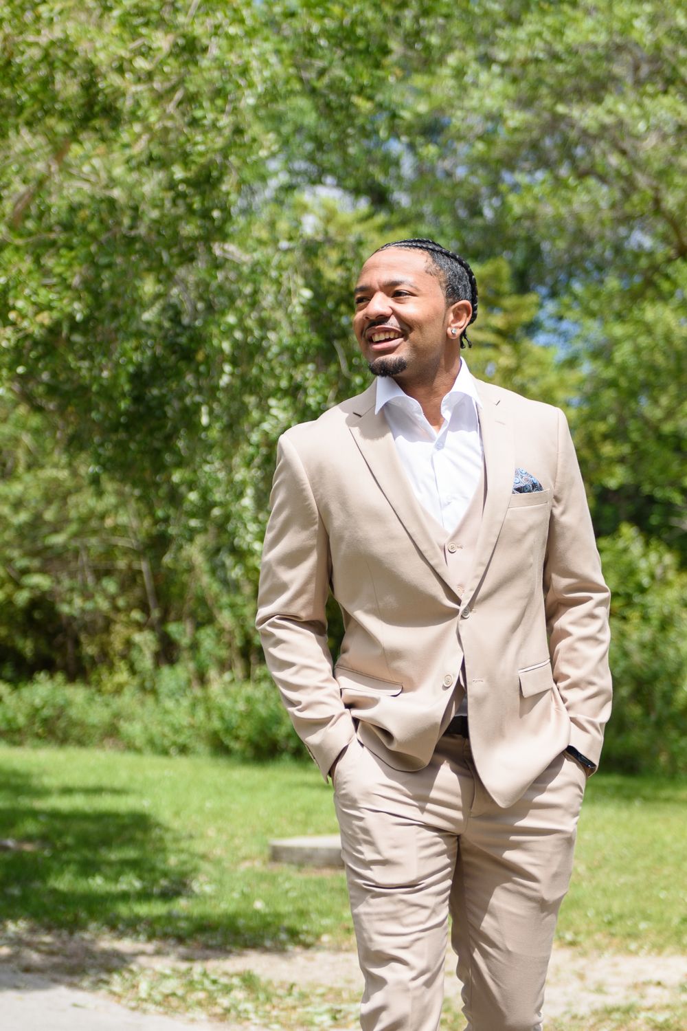 A person wearing a beige suit and white dress shirt stands outdoors smiling on a sunny day with greenery in the background.