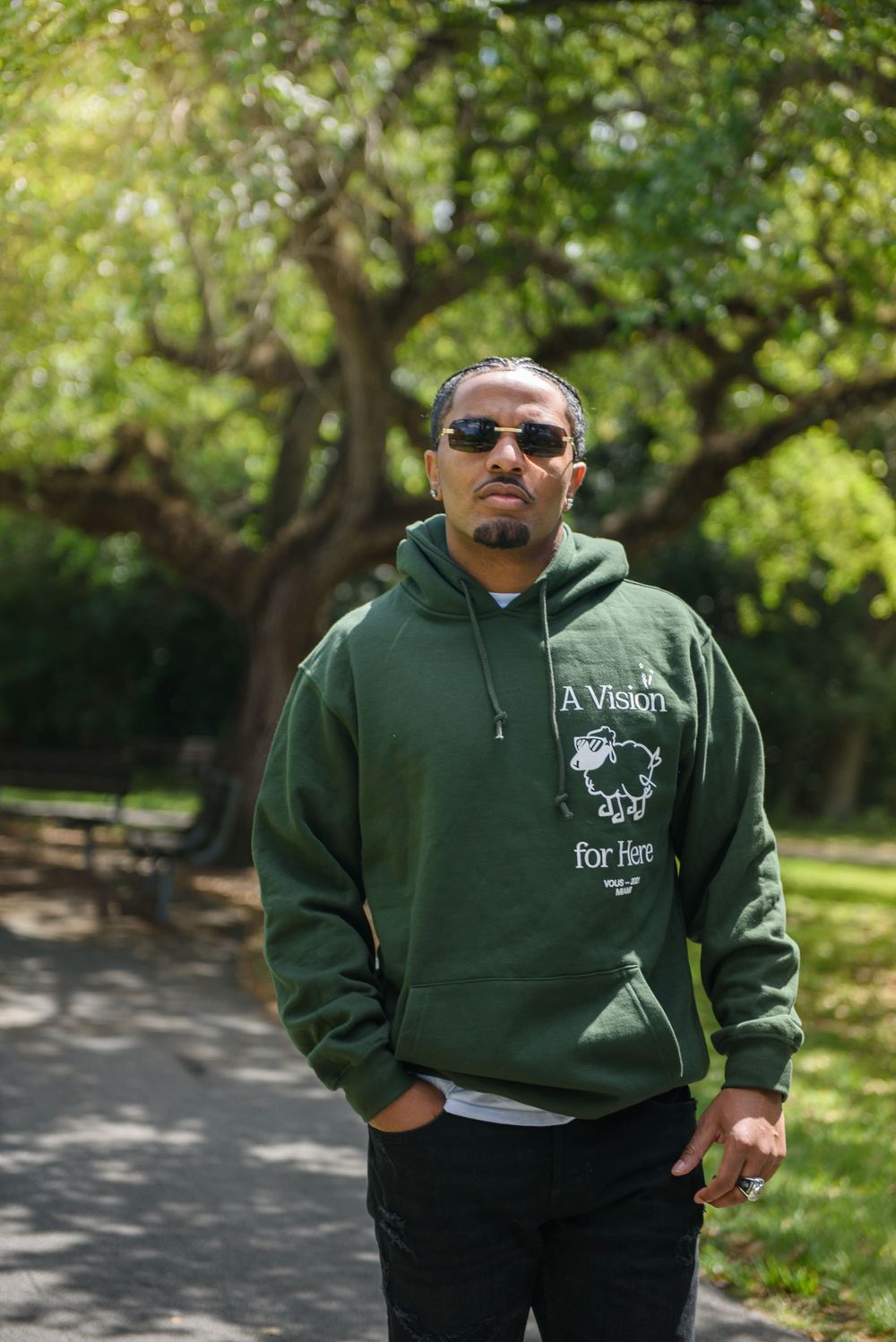 Person wearing sunglasses and a green hoodie standing on a tree-lined path in a park setting.