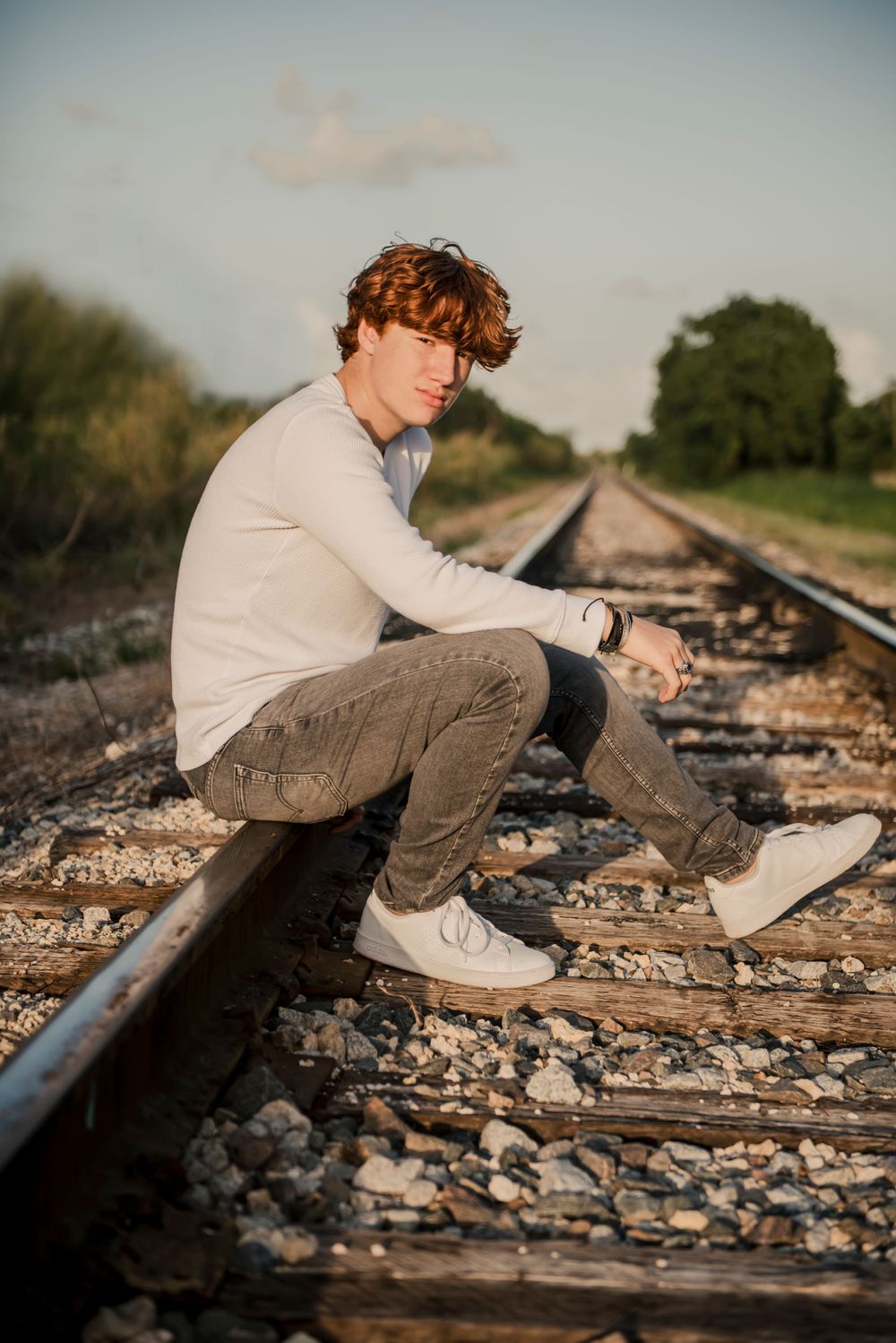 Senior sits contemplatively on railroad tracks wearing casual clothes at sunset.