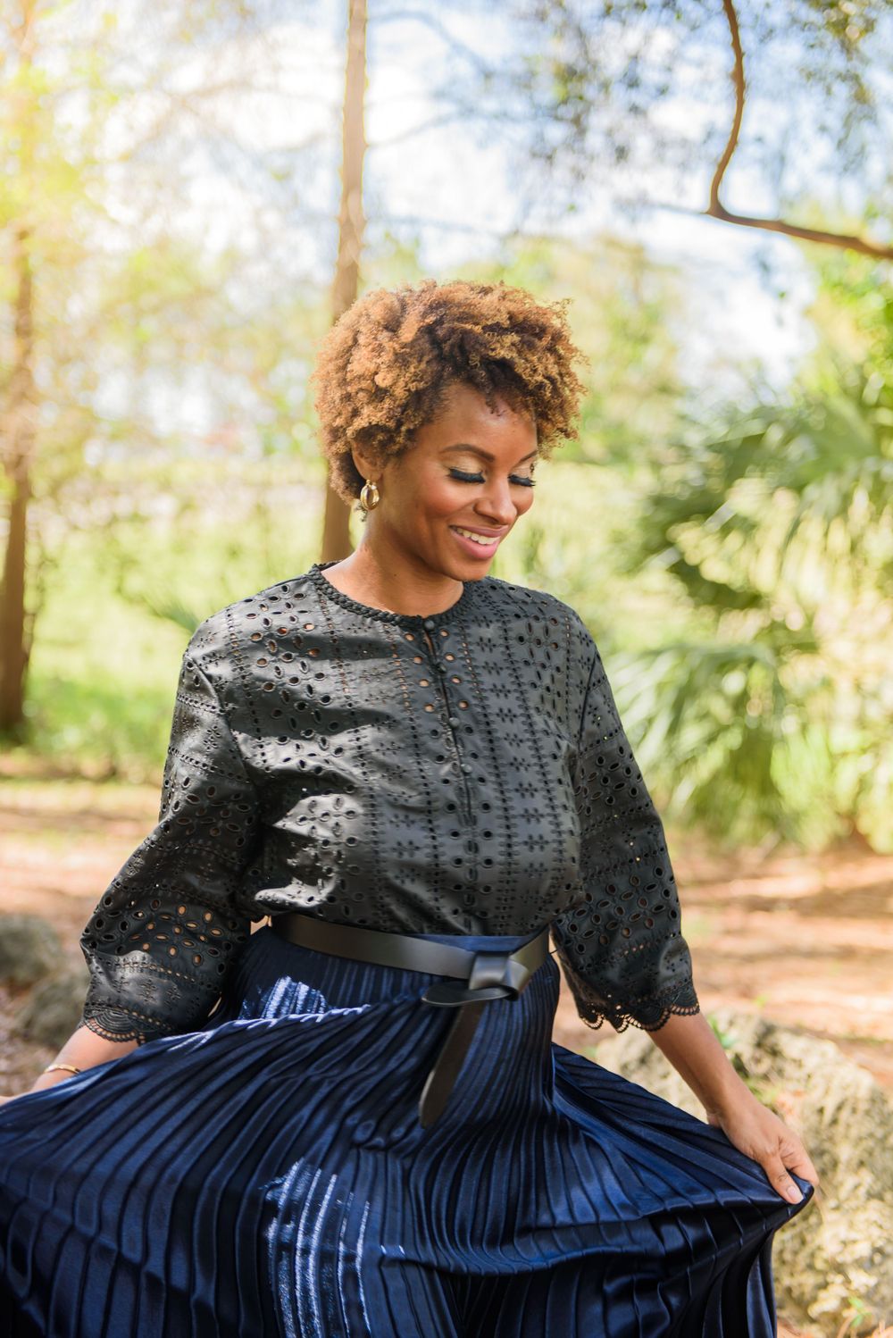 Stylish portrait in black eyelet top and navy pleated skirt among trees.