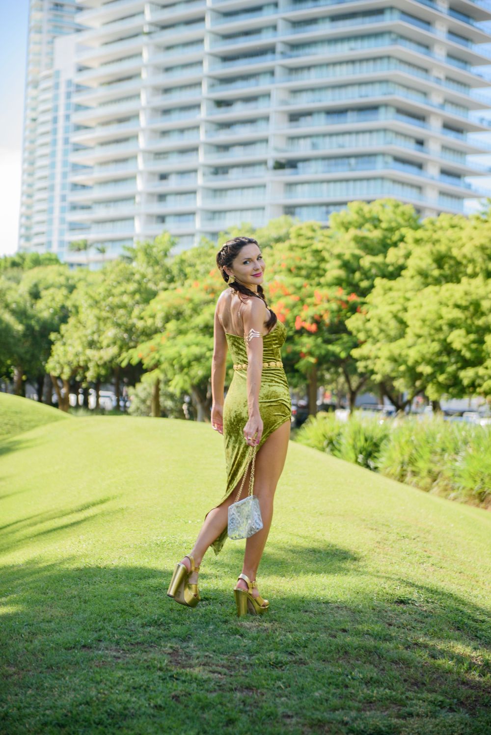 Fashion model in green dress poses on grassy hill with modern buildings in background.
