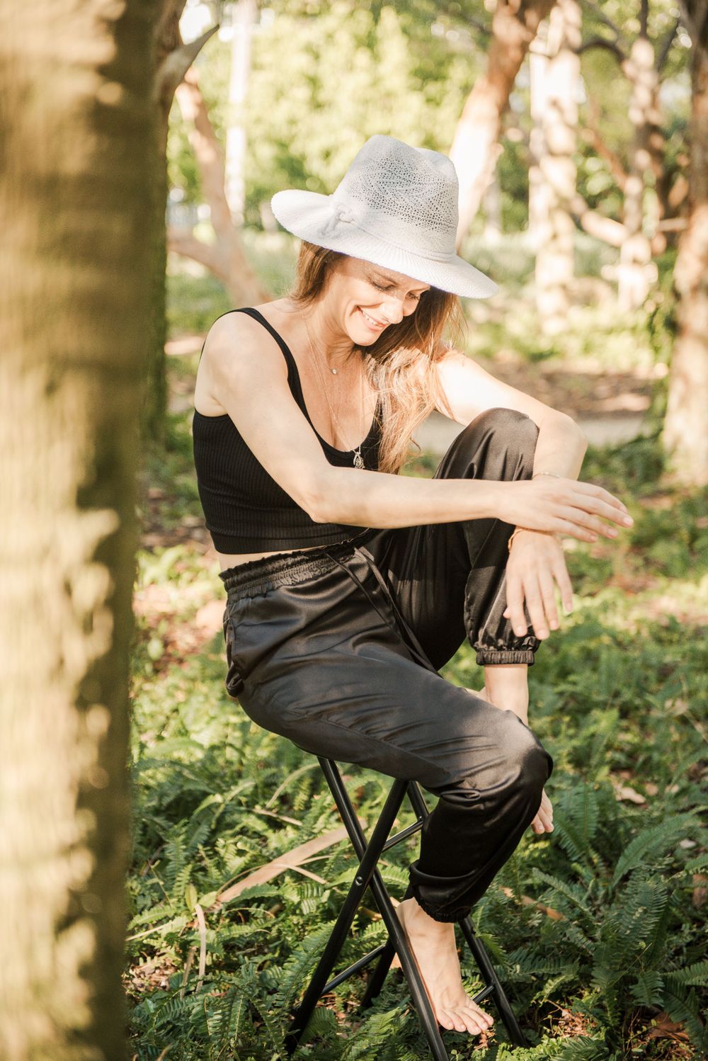 Artistic portrait of person in black outfit and hat sitting on stool in wooded setting.