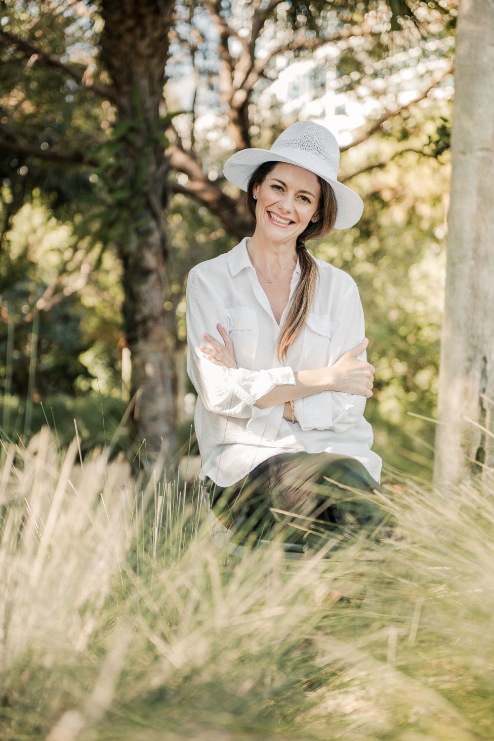 Stylish woman in white shirt and sun hat sits in tall grass during golden hour outdoors.