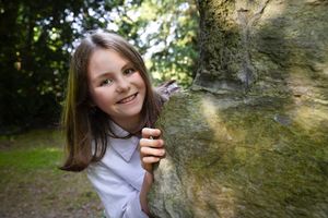 Person peeking playfully from behind large rock in wooded setting.