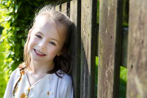 Smiling youth in white dress leaning against wooden fence in garden setting.