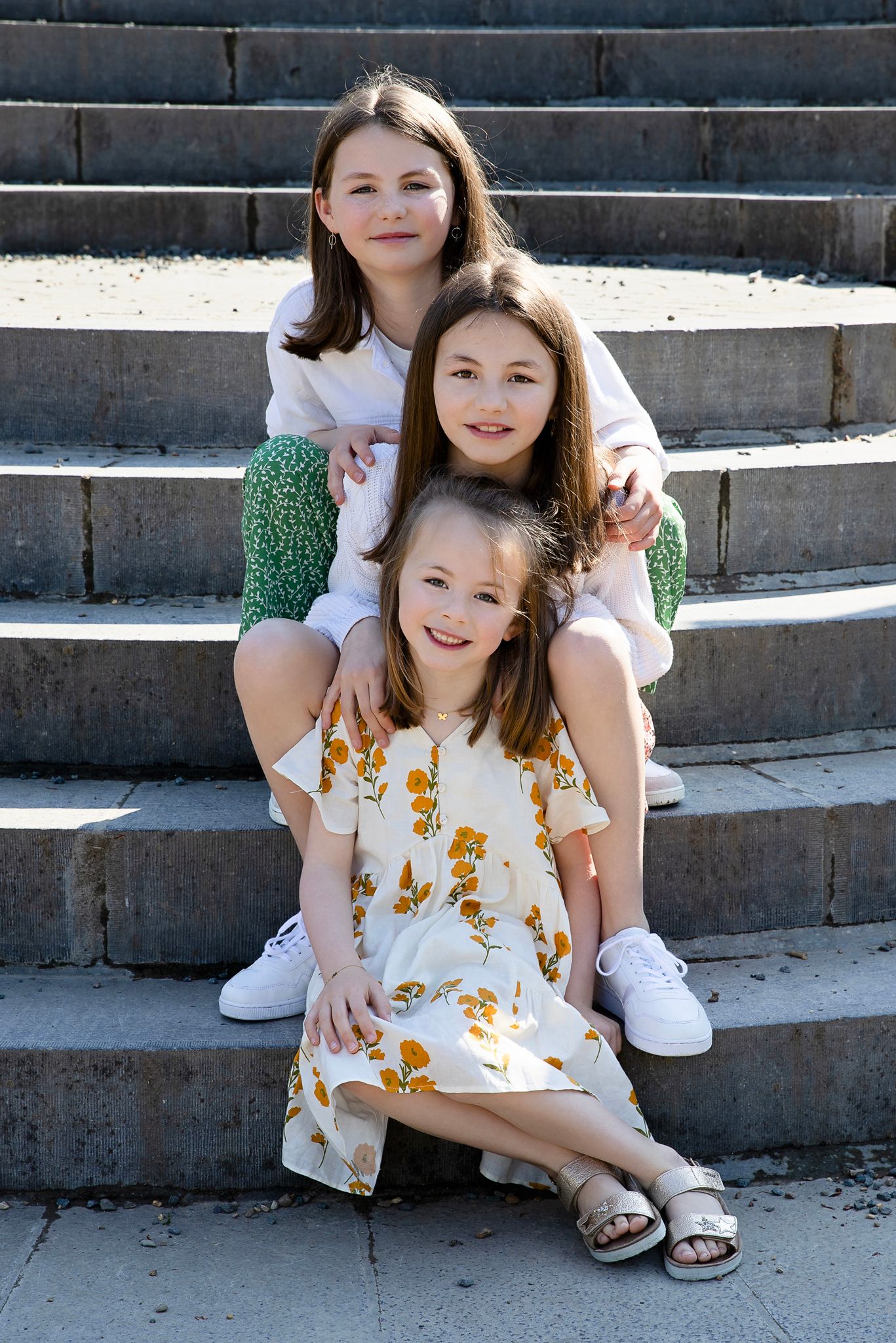 Three siblings sit casually on concrete steps outdoors with the oldest wearing green and youngest in a floral dress.