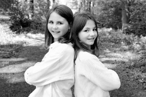 Black and white portrait of two young girls in sweaters standing back to back outdoors.