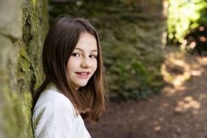 Portrait of young person in white sweater leaning against mossy stone wall in nature.