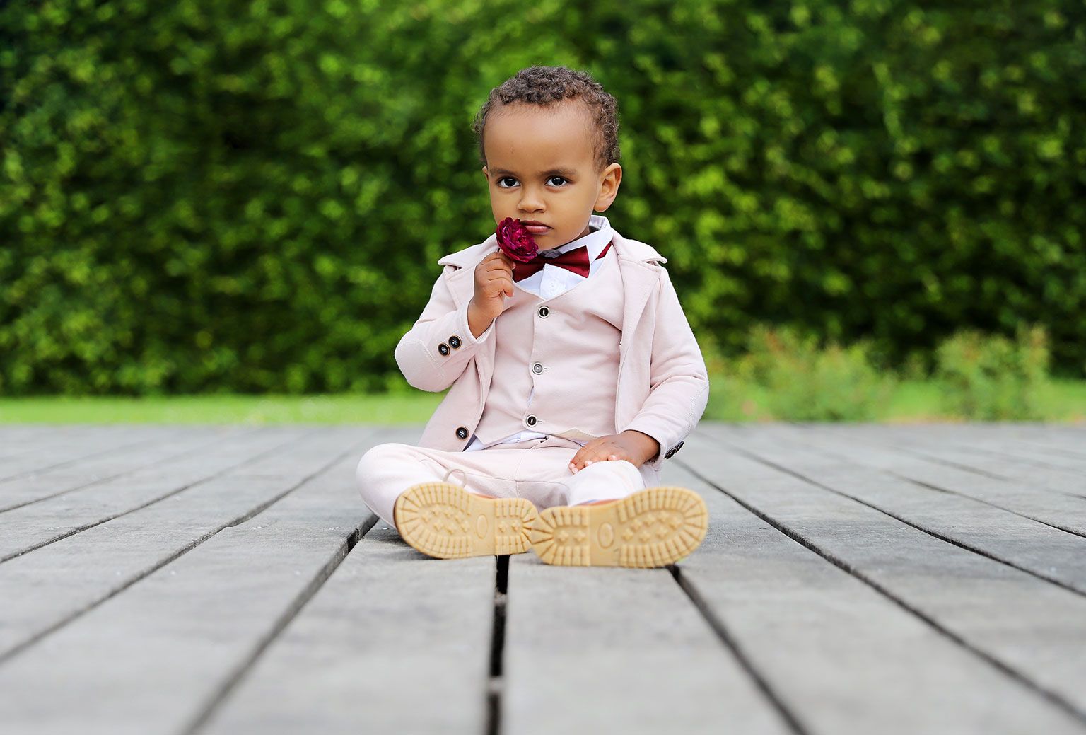 Young toddler in formal attire with bowtie sitting on wooden deck with greenery background.